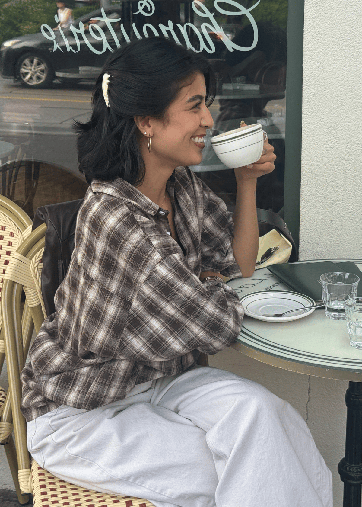 A woman with medium length hair in profile, sits at a table drinking a cup of coffee. She wears a light neutral hair comb in her hair to sweep her hair back from the side of her face. 