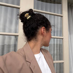 Woman with dark curly hair styled in a bun secured with a small beige-coloured hair clip wearing a beige blazer and white shirt, standing in front of a glass door.