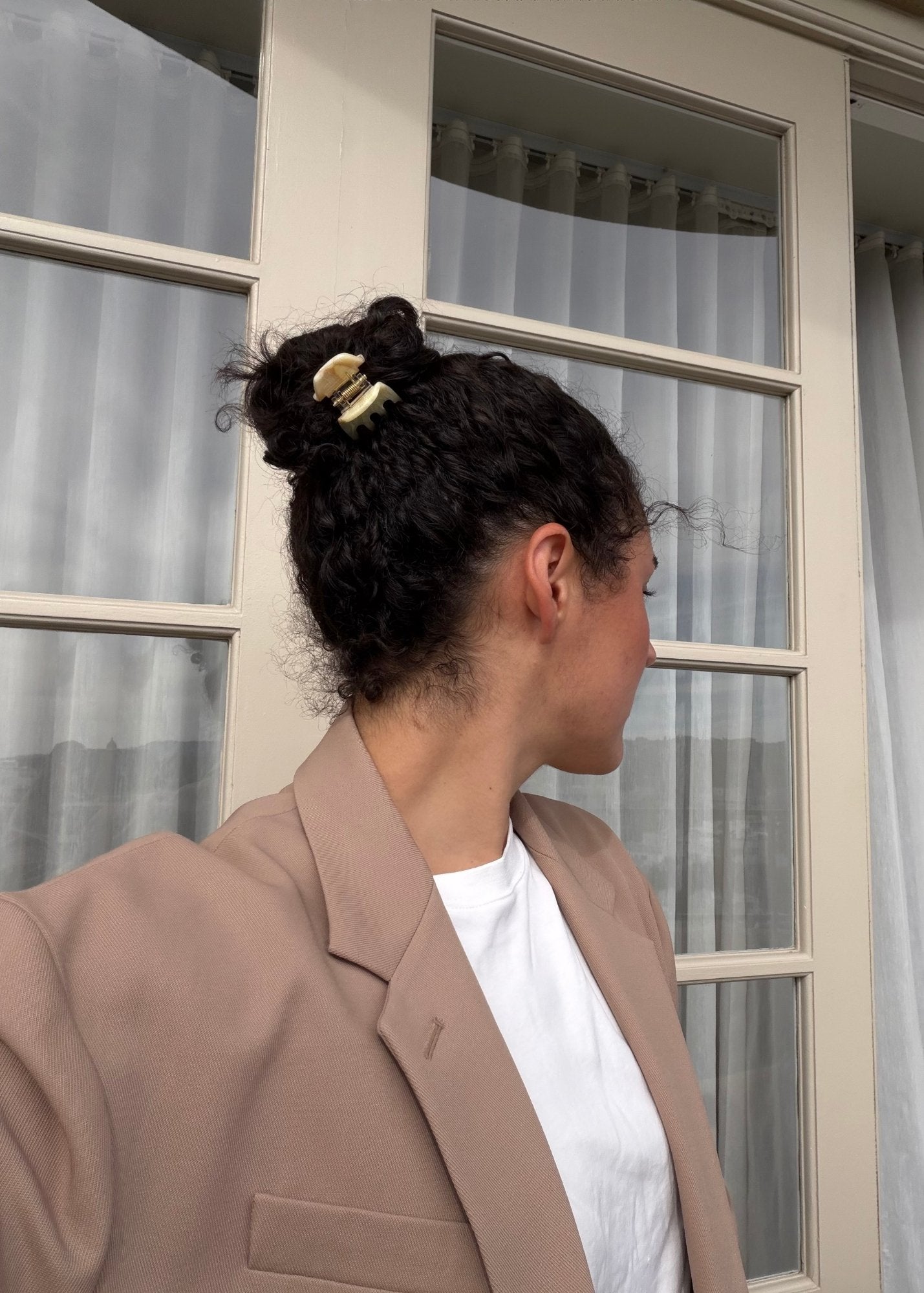 Woman with dark curly hair styled in a bun secured with a small beige-coloured hair clip wearing a beige blazer and white shirt, standing in front of a glass door.