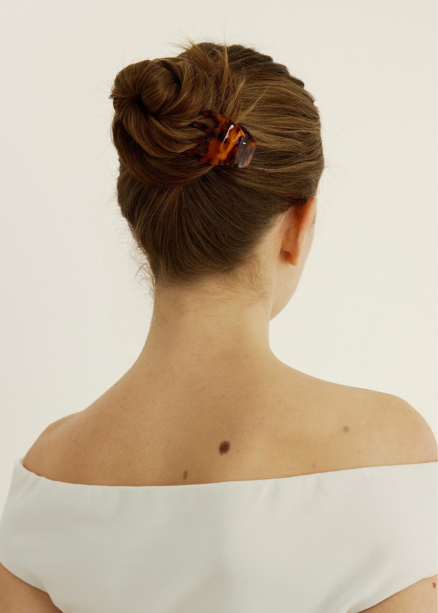 Back view of a person with a neatly styled bun secured with a small tortoise-coloured hair clip wearing a white off-shoulder top on a light background