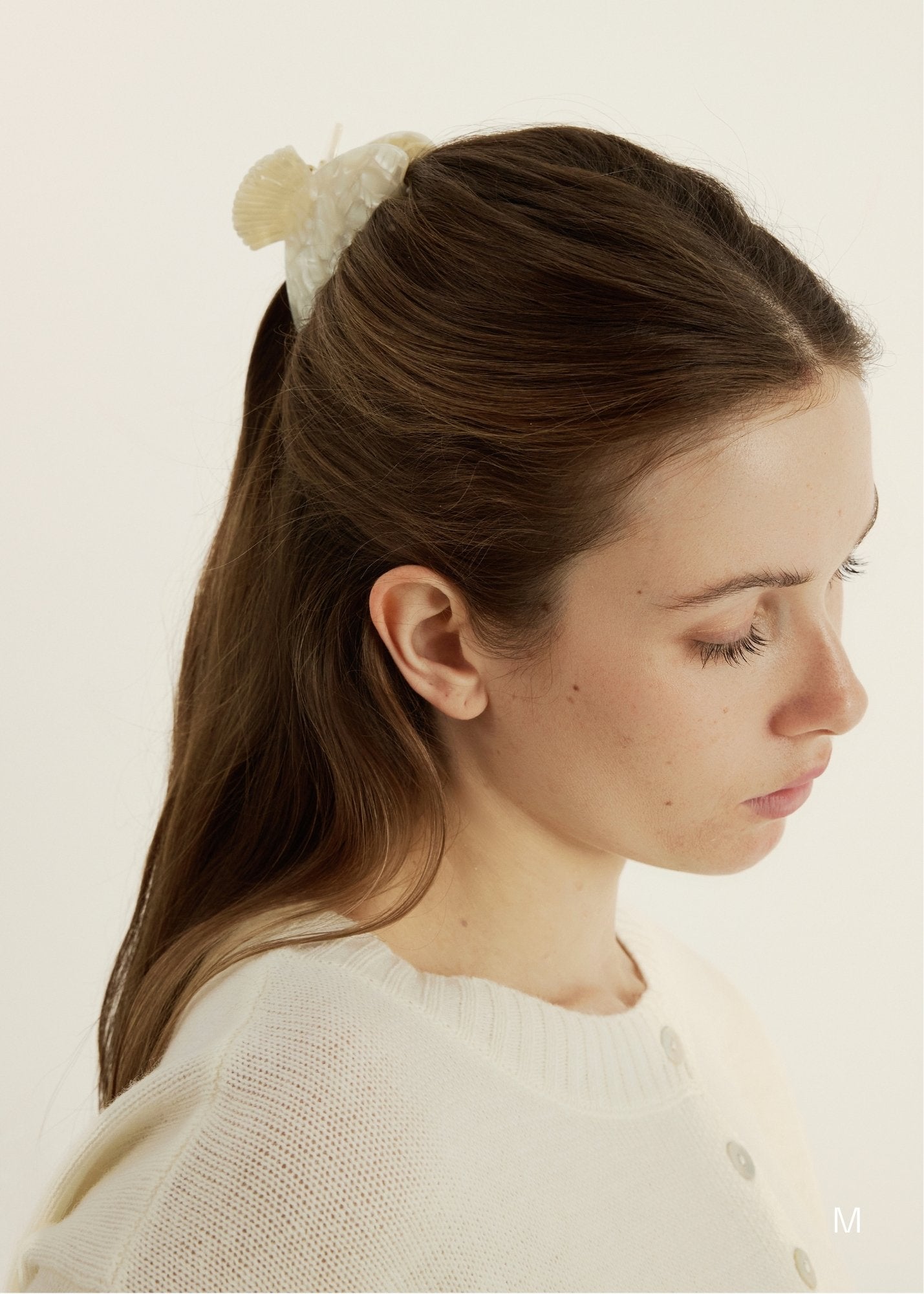 Woman with a half-up, half-down hairstyle wearing a shell inspired claw clip and a white sweater against a beige background