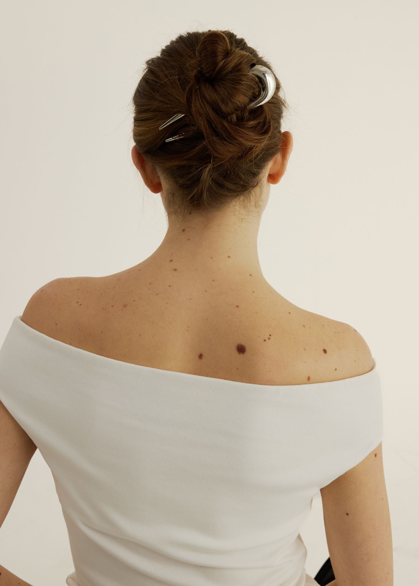 Back view of a woman wearing an off-shoulder white top with a plain background with her hair styled in an updo secured with the French Pin in Silver