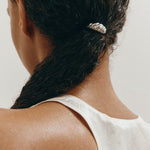 Woman with dark curly hair tied in a ponytail styled with a decorative croissant-shaped silver hair accessory against a neutral background
