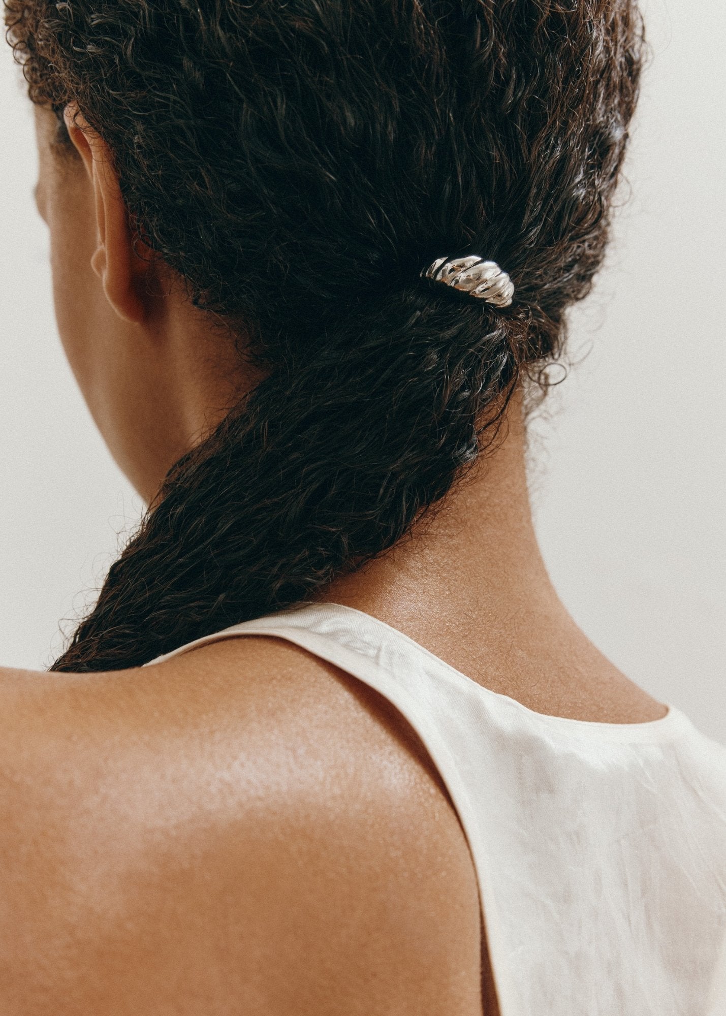 Woman with dark curly hair tied in a ponytail styled with a decorative croissant-shaped silver hair accessory against a neutral background