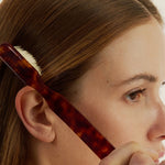 Woman using a small tortoise-coloured brush to style their hair against a neutral background