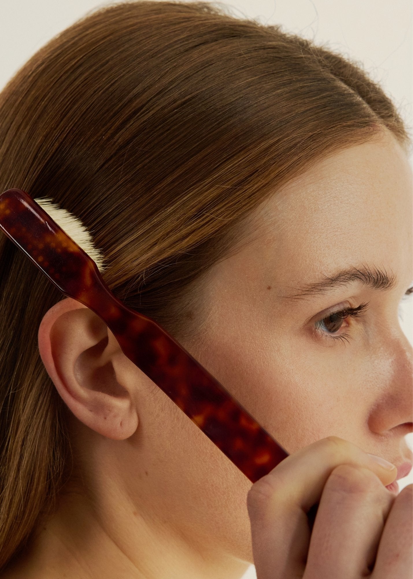 Woman using a small tortoise-coloured brush to style their hair against a neutral background