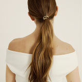 Woman with long brown hair in a ponytail hair cuff wearing an off-shoulder white top on a plain background