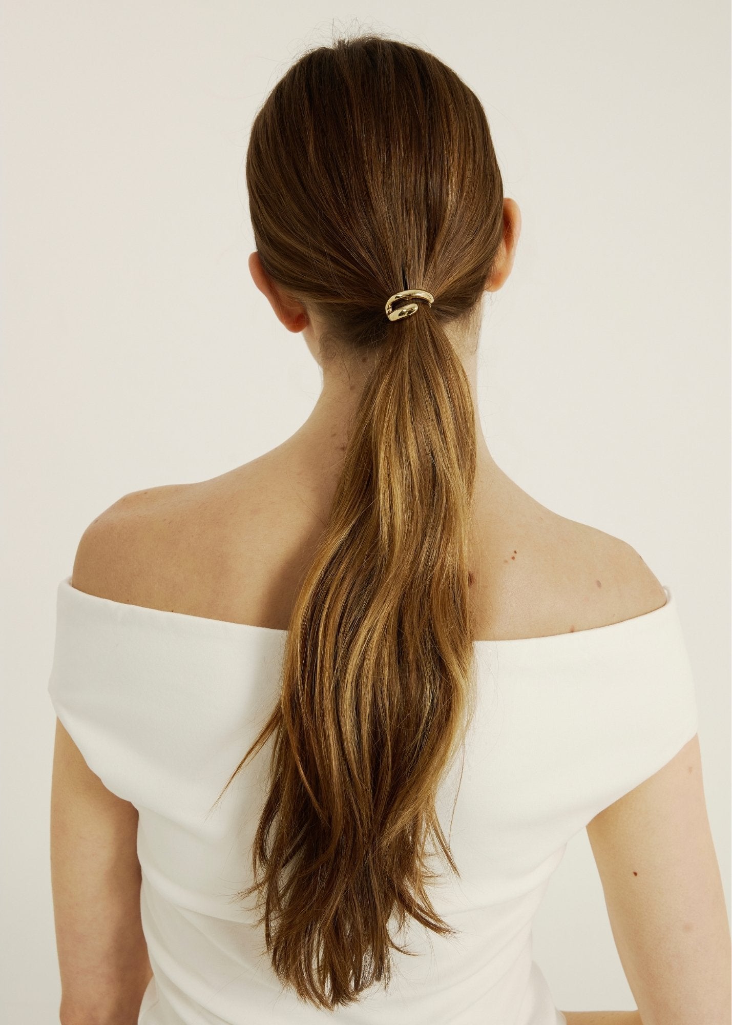 Woman with long brown hair in a ponytail hair cuff wearing an off-shoulder white top on a plain background