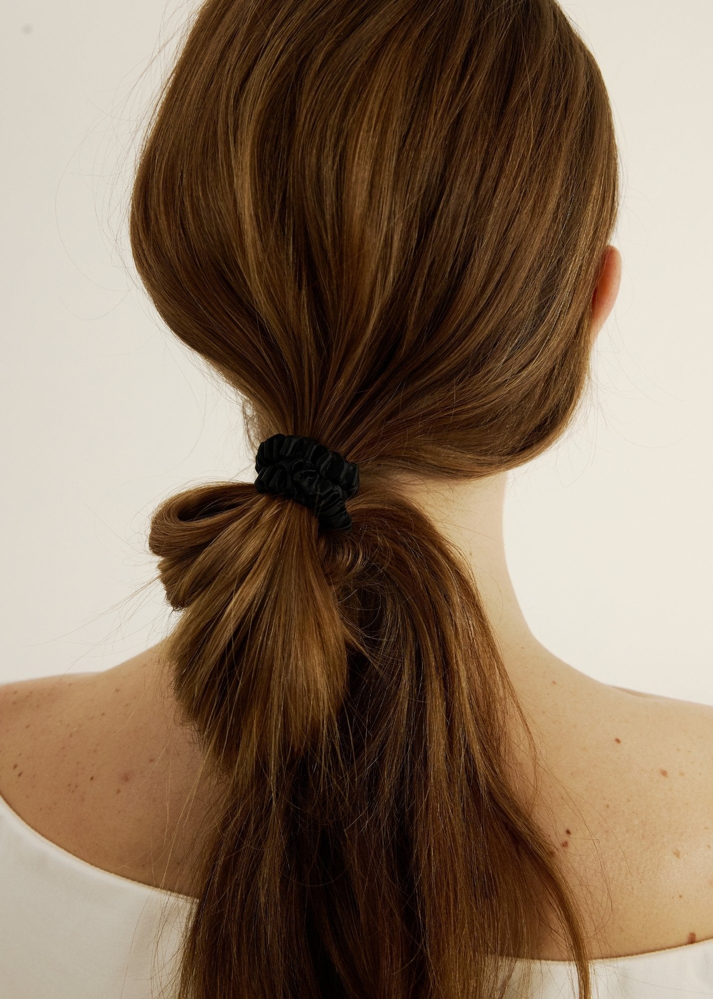 Back view of a person with long brown hair tied in a ponytail with a black scrunchie on a light background