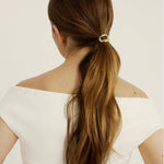 Woman with long brown hair styled in a ponytail wearing a hair cuff with a white off-shoulder top on a light background