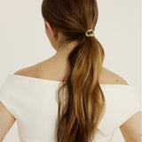 Woman with long brown hair styled in a ponytail wearing a hair cuff with a white off-shoulder top on a light background