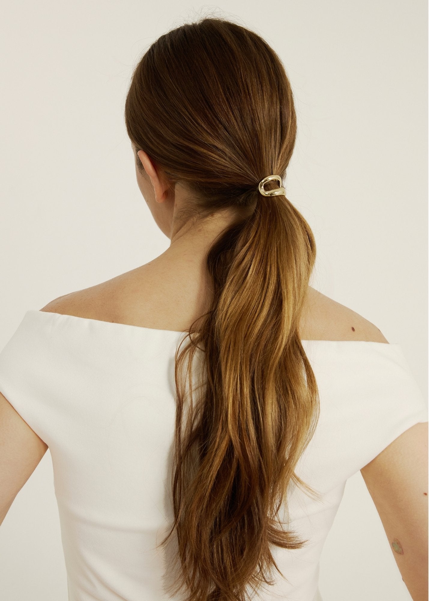 Woman with long brown hair styled in a ponytail wearing a hair cuff with a white off-shoulder top on a light background
