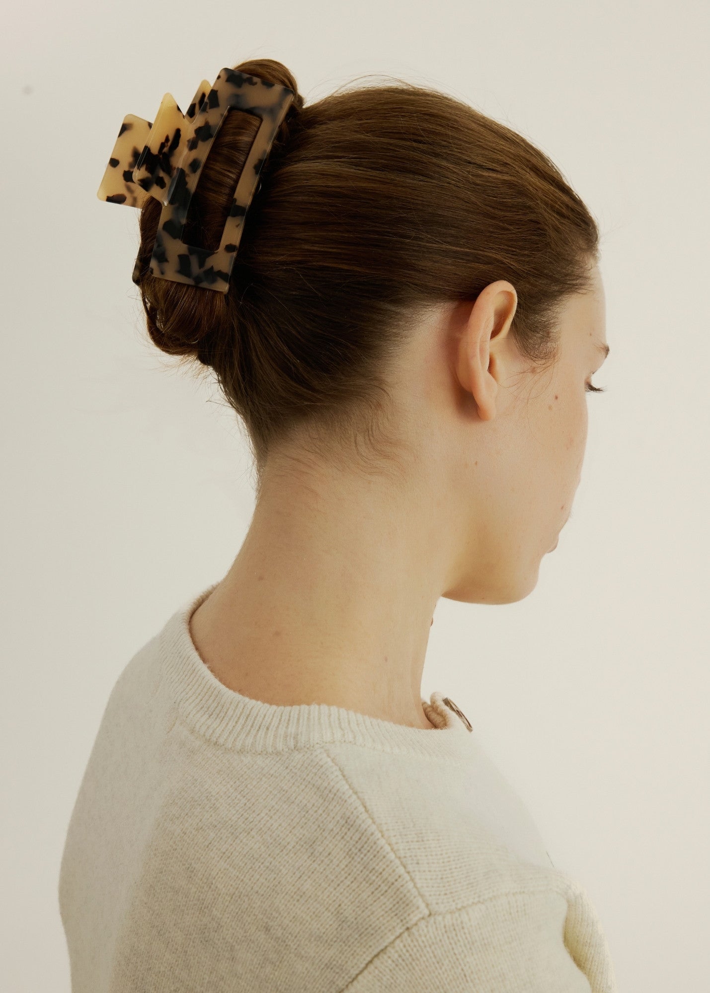 Close up view of a woman with straight, brown hair styled in an up do secured with a large, rectangular shaped, light tortoise hair claw clip against a white background.