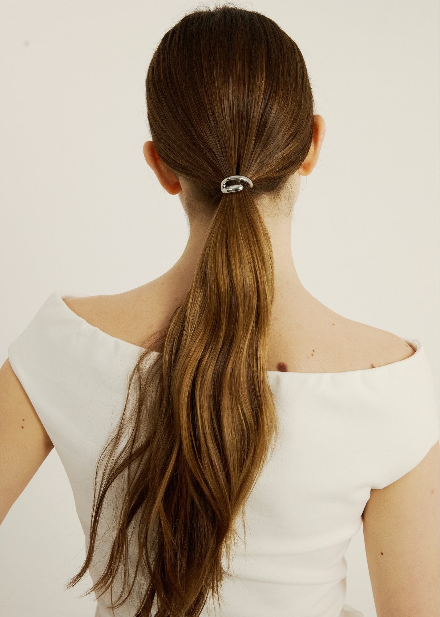 Woman with long brown hair tied in a ponytail hair cuff wearing a white top on a light background