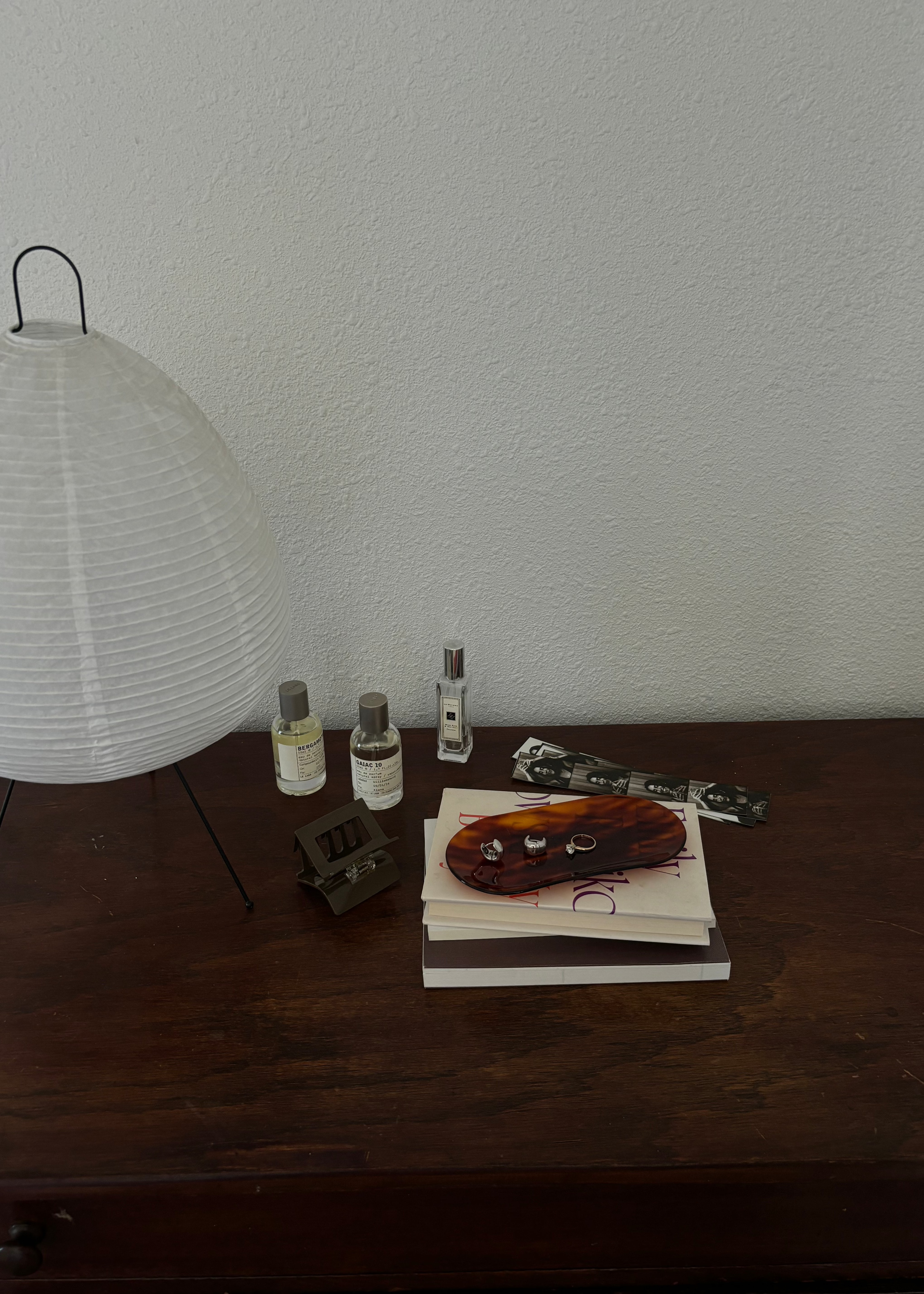 Photo of top of a wooden dresser with a white lamp, 3 perfume bottles, a large brown hair claw clip, a pile of books with a tortoise coloured tray with 3 silver rings on it.
